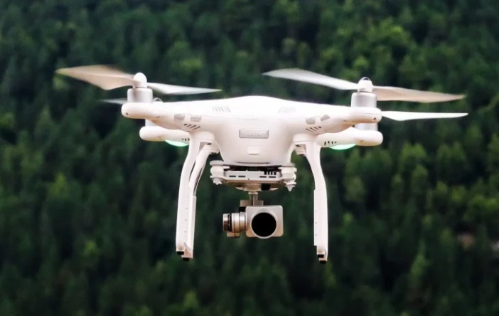 A white drone with a camera flying outdoors against a blurred green background.